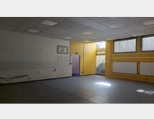 Ein leerer Innenraum mit gelben und weißen Wänden, Fliesenboden und einer Rasterdecke mit eingebauten Leuchten. An der Wand befinden sich Heizkörper und ein Notausgang führt zu einer Treppe im Außenbereich.