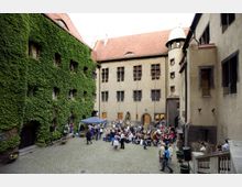 Ein historischer Innenhof mit Kopfsteinpflaster, umgeben von Gebäuden mit hellen Fassaden, von denen eines mit dichtem Efeu bewachsen ist. In der Mitte des Hofes befindet sich eine Versammlung von Menschen, teilweise unter einem Pavillon, während Statuen und Fenster die architektonischen Details ergänzen.