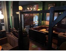 Gemütlicher Pub-Innenraum mit dunklen Holztrennwänden und Sitznischen, Holzbänken und Tischen sowie grünen Wänden mit gerahmten Schildern. Warme Hängelampen, ein Fenster mit Jalousien und dekorative Elemente wie Geländer und Fass ergänzen die rustikale Atmosphäre.