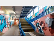 Ein Bahnhofstunnel mit Treppenaufgang, flankiert von Wänden, die mit farbenfrohen Graffitis bemalt sind. Auf der rechten Seite führen blaue Geländer und gelb markierte Treppenstufen nach oben, während das Licht durch große Fenster einfällt.