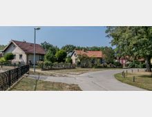 Straßenszene in einem Wohngebiet mit kleinen Einfamilienhäusern, die rote Ziegeldächer und Vorgärten mit Pflanzen aufweisen. Ein gepflasterter Weg schlängelt sich durch die Umgebung, begleitet von Rasenflächen, Bäumen und einem Straßenlaternenpfahl.