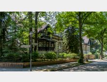 Eine historische Wohnstraße mit einem eleganten Fachwerkhaus im Jugendstil, umgeben von hohen Bäumen und gepflegtem Grün. Straßenschilder für „Salzachstraße“ und „Bergengruenstraße“ sowie eine alte Straßenlaterne sind sichtbar, mit Kopfsteinpflaster am Gehweg.