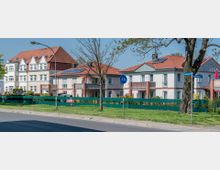 Straßenszene mit zweistöckigen Wohnhäusern, die rote Satteldächer und Balkone haben; im Vordergrund ein Gehweg, eine Straße und grüne Wiesen mit einem Baum. Ein Verkehrsschild für einen Radweg und Straßenschilder befinden sich ebenfalls im Bild.