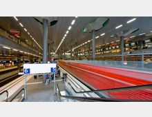 Großer, moderner Bahnhof mit mehreren Ebenen und Bahnsteigen. Im Vordergrund sind die Gleise 5 und 6, eine rote, schnell fahrende Zugverbindung sowie einige Reisende sichtbar, umgeben von glänzenden Metallstrukturen und künstlichem Licht.