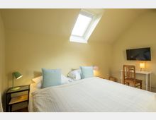 Schlafzimmer mit gelben Wänden, einem Doppelbett mit pastellfarbenen Kissen, einem Dachfenster, Nachttischen mit Lampen und einem kleinen Bereich mit Schreibtisch, Stuhl und Wandfernseher.