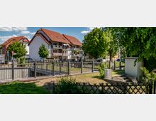 Mehrfamilienhäuser mit roten Schrägdächern und Balkonen stehen im Hintergrund, umgeben von Bäumen. Im Vordergrund befindet sich eine kleine Brücke mit einem Geländer, technische Anlagen und eine grüne Wiese.