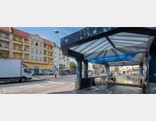 U-Bahn-Eingang "Franz-Neumann-Platz (Am Schäfersee)" an einer belebten Straßenkreuzung, mit einem verglasten Überdachungsbau und Rolltreppen, umgeben von gelben Wohnhäusern, Geschäften und vorbei fahrenden Fahrzeugen.