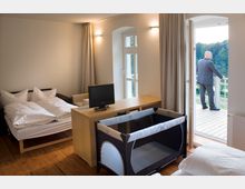 Ein modernes Hotelzimmer mit Doppelbett, Holzboden, Schreibtisch, Fernseher und einem aufgestellten Babybett. Eine geöffnete Balkontür führt zu einem Balkon mit Blick auf eine grüne Landschaft, auf dem ein Mann steht.