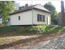Einfache, eingeschossige Haus mit weißer Fassade, schwarzen Fensterläden und rotem Satteldach, umgeben von Bäumen. Im Vordergrund ist ein leicht abschüssiges Gelände mit gepflastertem Weg und Laub zu sehen.
