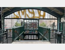 Ein U-Bahnhof mit grün lackierter Metallkonstruktion und einem Treppenabgang nach unten. Im Hintergrund sind historische Gebäude, Bäume und Stadtverkehr zu sehen; die Fensterscheiben im oberen Bereich sind teils mit Graffiti bedeckt.