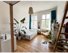 Helles Schlafzimmer mit Holzboden, einem Bett, großen Zimmerpflanzen und hohen Fenstern mit blauen Vorhängen. Rechts steht eine Holzleiter, die zu einer oberen Ebene führt, neben einem Regal und einem gelben Sessel.