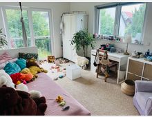 Ein helles Kinderzimmer mit Teppichboden, einem Tagesbett voller Kuscheltiere, einem Schreibtisch mit Stuhl und Dekorationen sowie Spielzeug, das auf dem Boden verteilt ist. Große Fenster lassen Tageslicht herein und bieten Ausblick auf einen Garten mit Bäumen.