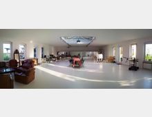 Großer, hell erleuchteter Raum mit weißem Boden und vielen Fenstern, die natürliches Licht hereinlassen. Der Raum enthält verschiedene Sitzmöbel, darunter braune Ledersessel und einen Tisch mit Stühlen in der Mitte; eine weiße Kunststoffstatue einer Kuh steht prominent auf einer Seite, während technische Geräte und Möbel im Hintergrund sichtbar sind.