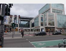 Straßenszene vor einem modernen Einkaufszentrum in Berlin, bestehend aus zwei Gebäuden mit großen Glasfassaden. Zu sehen sind Geschäfte wie "Karstadt" und "Saturn", Passanten auf dem Gehweg und ein Radweg im Vordergrund.