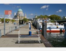 Uferpromenade mit einem Anlegesteg, auf dem rechts ein Ausflugsschiff mit der Aufschrift „Cecilienhof Potsdam D“ liegt; im Hintergrund ist die Nikolaikirche mit ihrer großen Kuppel sowie weitere historische Gebäude und Palmen auf dem Platz zu sehen. Der Himmel ist blau mit wenigen Wolken.