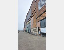 Industriedenkmal mit Backsteinfassade und großen Fenstern, rechts vorne ein Wandmosaik sowie Hinweistafeln. Der Bereich davor besteht aus einem breiten Betonweg, der an eine offene Freifläche grenzt.