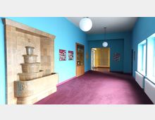 Flur mit hellblauen Wänden und dunkelrotem Teppichboden, links ein Wandbrunnen aus beigefarbenen Kacheln, daneben Vitrinen und Informationstafeln. Große Fenster auf der rechten Seite und runde Deckenleuchten sorgen für Beleuchtung.