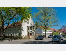 Straßenszene in einer Wohngegend mit historischen weißen Häusern und roten Ziegeldächern, flankiert von großen Bäumen. Am Straßenrand stehen geparkte Autos, und im Hintergrund sind weitere Häuser sowie blühende Bäume zu sehen.