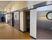 Flur mit gelblich-braunem Boden, weiß gestrichenen Wänden und mehreren großen, silbernen Schiebetüren mit runden Fenstern. Eine der Türen ist geöffnet und zeigt ein dahinterliegendes Badezimmer mit Fliesen und einem Waschbecken. Links im Bild steht eine Person nahe einer Glastürkonstruktion.