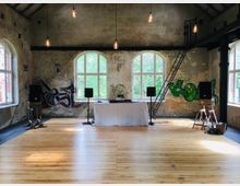 Ein rustikaler, loftartiger Raum mit hohen Decken, sichtbaren Holzbalken und großen, gewölbten Fenstern, durch die grünes Außengelände sichtbar ist. Im Vordergrund eine Holzfußbodenfläche, eine DJ-Konsole mit weißer Tischdecke, Lautsprecherboxen auf Ständern und Graffiti an den Wänden.