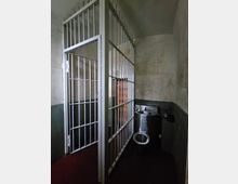 Innenansicht einer Gefängniszelle mit weiß gestrichenen Gittertüren, schlichten Wänden mit Abnutzungsspuren und einer Edelstahltoilette mit integriertem Waschbecken. Die Umgebung ist schlicht und funktional gestaltet.