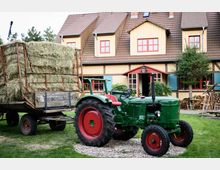 Traktor mit angehängtem Heuwagen steht auf einer Wiese vor einem Fachwerkhaus mit roten Fensterrahmen und blauen Fensterläden. Im Hintergrund sind Holzbündel und ein kleiner Baum sichtbar.