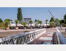 Moderne Außenanlage mit einem Cafébereich, Palmen in großen Pflanzkübeln und weißen Sonnenschirmen, im Hintergrund ein Gebäude mit Glasfassade. Im Vordergrund führt eine hell gestrichene Fußgängerbrücke über einen kleinen Wasserlauf, rechts eine schräg aufragende Metallkonstruktion.