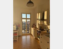 Sunlit residential kitchen with white shaker-style cabinets, wooden countertops, a white tile backsplash, and a black range hood over the stove. A tall glass door with divided panes opens to a small balcony, and a black pendant light hangs above wide wooden floorboards.