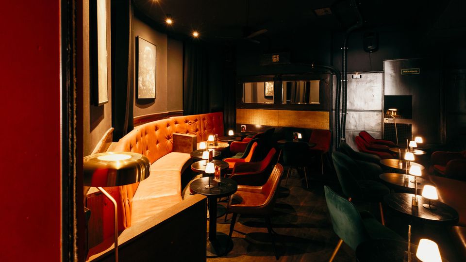 Gedimmter Lounge- oder Bar-Innenraum mit dunklen Wänden, holzgemustertem Boden und einer langen, gepolsterten Sitzbank in Orange. Viele kleine runde Tische mit Tischlampen sowie Sessel in Rot- und Grüntönen stehen in Reihen; im Hintergrund sind Spiegel- und Metallwandflächen sowie eine Tür zu sehen.