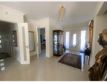 Eingangsbereich eines Wohnhauses mit hellem Fliesenboden, einer verglasten Vitrine, einem orientalischen Teppich und einer goldfarbenen Statue. Im Hintergrund befinden sich eine Doppeltür mit Milchglas und mehrere angrenzende Räume mit offenen Türen.