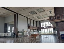 Innenansicht einer großen Halle mit hoher Decke und rechteckigen Kassettenelementen. Die Wände bestehen aus einer Kombination aus Backstein und hellen Flächen; auf der rechten Seite befindet sich eine umlaufende Holzbalustrade, darunter große Fensterfronten mit Gitterstruktur, die viel Tageslicht hineinlassen. Auf dem Boden liegt ein hell-dunkel kariertes Muster, und der Raum ist mit Schautafeln, Vitrinen und Ausstellungsstücken bestückt.