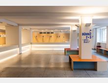 Heller, moderner Innenraum einer Veranstaltungs- oder Kulturstätte mit breitem Tresen vor einer Holzverkleidung, weiß gestrichenen Stützen und gefliestem Boden. Rechts stehen farbige Sitzbänke um eine Säule mit Wegweiser-Aufschrift „BÜHNE 2+3“, daneben große Fensterfront.