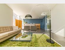 Heller Wohnraum mit modernen Möbeln, darunter ein beiges Sofa, zwei schwarze Sessel und ein Glastisch auf einem grünen Teppich mit Schriftzügen. Im Hintergrund eine blaue Wand, ein Schreibtisch mit Pflanzen und eine große Bogenlampe im Vordergrund.