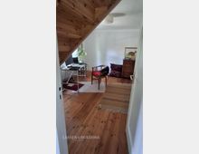 Blick in ein kleines Dachgeschosszimmer mit schräger Holzdecke und Holzdielenboden. Links steht ein einfacher Schreibtisch mit Monitor und Pflanze am Fenster, rechts eine Kommode mit Bild sowie Sitzkissen und ein Stuhl auf Teppichen.