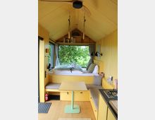 Innenansicht eines kleinen Tiny Houses mit holzverkleidetem Satteldach, eingebauter Schlafnische vor einem großen Fenster mit Blick ins Grüne und integrierten Regalen darüber. Vorne stehen ein kleiner Tisch, gepolsterte Sitzbänke und rechts eine kompakte Küchenzeile mit Kochfeld und Spüle.