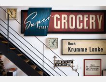 Innenraum mit Dekoration an einer weißen Wand, bestehend aus vintage Schildern mit Texten wie "Super Store Grocery" und "Nach Krumme Lanke" sowie zwei analogen Uhren. Im Vordergrund eine schwarze Metalltreppe mit Holzstufen.