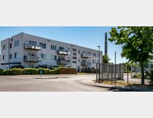 Mehrstöckiges Wohnhaus mit weißen Fassaden und Balkonen, umgeben von niedrigen Hecken. Im Vordergrund befindet sich eine Straße mit Bürgersteig und ein offenes Tor, im Hintergrund ein industrieähnlicher Schornstein sowie einige Bäume.