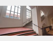 Treppenhaus in einem historischen Gebäude mit großen, hohen Fenstern, die viel Tageslicht hereinlassen. Der Bereich hat weiße Wände, rote Böden und schwarze Geländer an den Treppen, ergänzt durch elegante Rundbögen im Hintergrund.