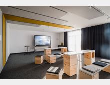 Moderner Schulungsraum mit modularen Holzmöbeln, darunter Sitzhocker und Tische, sowie einem großen interaktiven Bildschirm an der Wand. Der Raum hat schallabsorbierende Deckenelemente, grauen Teppichboden, große Fenster mit Vorhängen und wirkt hell durch natürliche Lichtquellen.