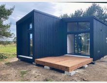 Modernes, kubisches Tiny House mit schwarzer Holzfassade und asymmetrischem Pultdach, davor eine Holzterrasse auf Betonfundamenten. Große bodentiefe Glasfronten und schmale Fenster, gelegen auf einer offenen Wiese mit Bäumen am Rand.
