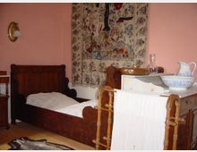 Ein historisches Schlafzimmer mit einem dunklen Holzbett, einer Kommode mit Waschutensilien wie einer Keramikschüssel und Krug sowie einem Handtuchhalter im Vordergrund. An der Wand hängt ein bunt bestickter Wandteppich, und der Raum ist in einem zartrosa Farbton gestrichen.