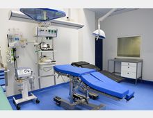 Moderner Operationssaal mit blau gepolstertem Operationstisch, medizinischen Geräten wie Beatmungsgeräten und Monitoren sowie Deckenlampen. Der Raum hat eine sterile Einrichtung mit weißen Wänden und blauem Bodenbelag.