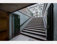Treppe in einem unterirdischen Fußgängertunnel, die nach oben zu einer Straße mit Altbaufassaden führt. Die Wände sind mit kleinen Fliesen verkleidet, und links befinden sich Gittertüren.