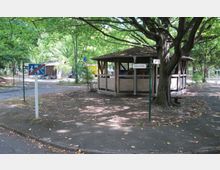 Eine kleine Holzhütte steht in einer parkähnlichen Umgebung unter einem großen Baum. Im Vordergrund befinden sich ein Straßenschild, das ein Wohngebietende anzeigt, sowie ein Schild mit der Aufschrift "Gubener Straße".