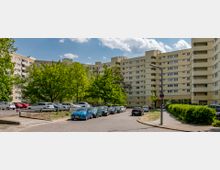 Mehrstöckiges Wohngebäude mit hellgelber Fassade, umgeben von geparkten Autos und grünen Bäumen. Im Vordergrund eine asphaltierte Straße, daneben Gehwege und Büsche.