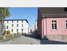 Straßenszene in einer historischen Stadt mit gepflasterter Straße, flankiert von weißen und rosa Gebäuden mit klassischen Fenstern. Im Hintergrund ist ein Turm mit grünlicher Kuppel zu sehen, links stehen Bäume und eine Parkbank.