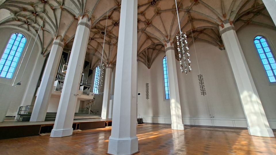 Innenraum einer hohen gotischen Hallenkirche mit weißen Pfeilern, Kreuzrippengewölbe und großen, schmalen Rundbogenfenstern. Im Hintergrund ist eine Orgel auf einer Empore zu sehen, von der Decke hängen mehrere moderne Leuchter über dem Holzfußboden.