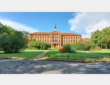 Backsteingebäude im neogotischen Stil mit einem Mittelgiebel, einem Uhrturm und zahlreichen hohen Fenstern, umgeben von einer gepflegten Parkanlage mit Blumenbeeten, Rasenflächen und Gehwegen. Im Vordergrund ist eine Straße zu sehen.