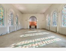 Heller Innenraum einer historischen Kapelle mit hohen, bunten Glasfenstern an den Seitenwänden. Der Raum hat einen gemusterten Steinboden, weiß gestrichene Wände mit Rundbögen und im Hintergrund einen zentralen Altarbereich unter einer kleinen Kuppel.