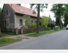 Eine alte Wohnhausfassade mit teils verwittertem Putz und roten Dachziegeln, umgeben von Bäumen und Büschen. Vor dem Haus befindet sich ein roter Holzzaun, der an einen Gehweg und eine asphaltierte Straße grenzt.